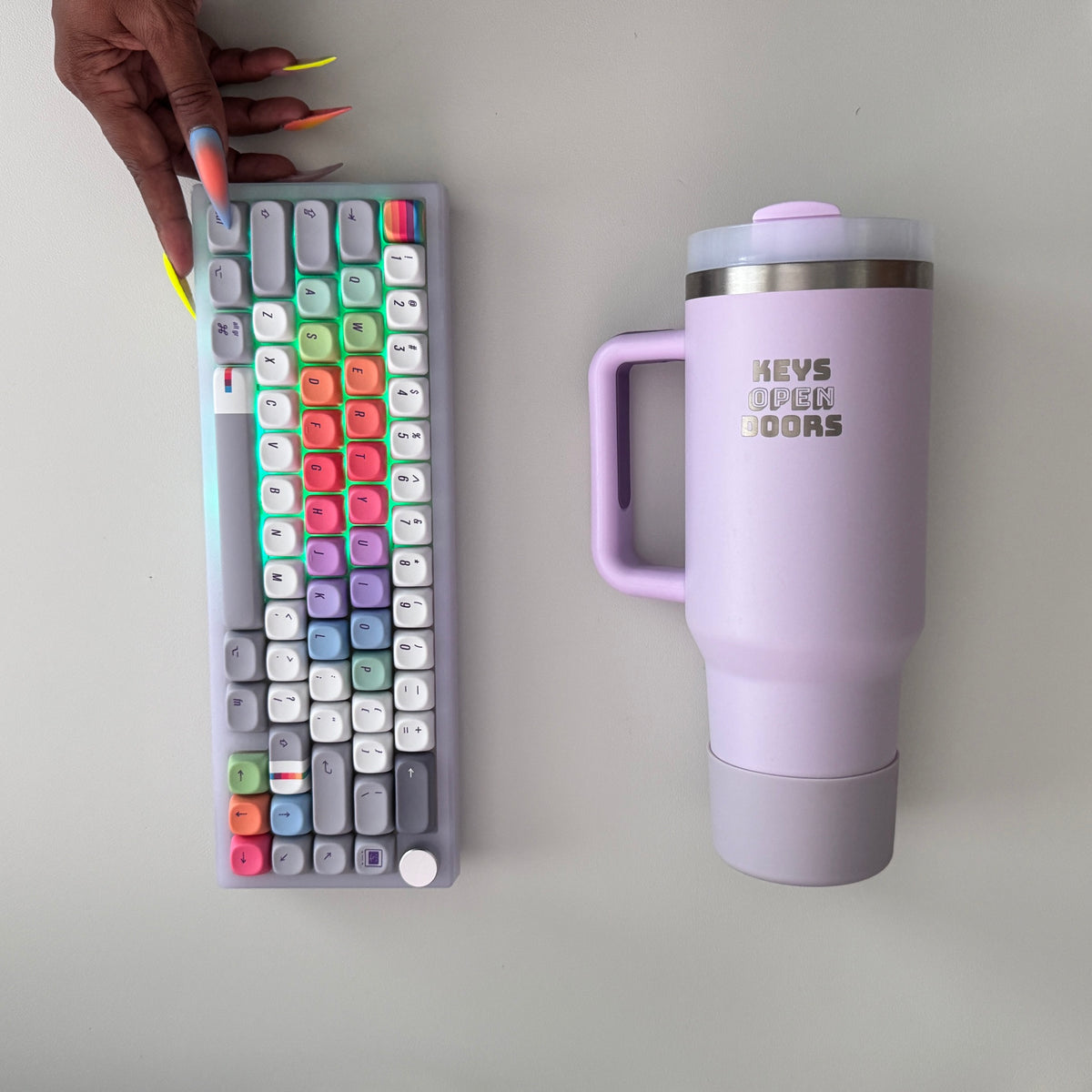 Purple Frost keyboard for Mac with green backlight and rainbow keycaps beside the lilac Keys Open Doors tumbler