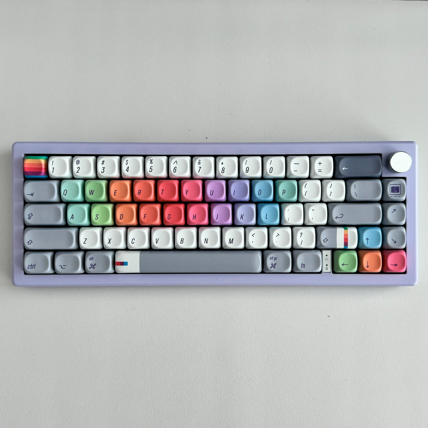 A compact mechanical keyboard for Mac with a lavender-purple chassis and a full set of pastel-colored keycaps arranged in rainbow rows, photographed from directly above on a neutral light gray background.