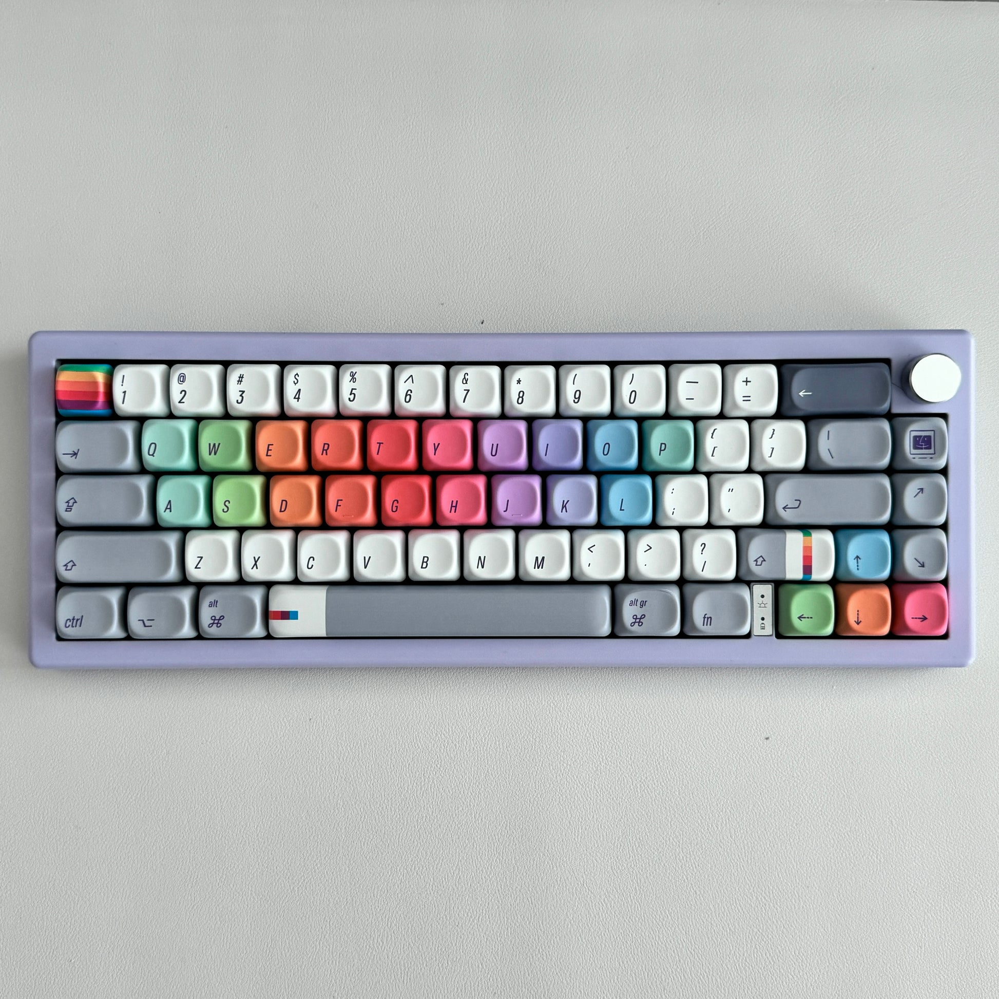 A compact mechanical keyboard for Mac with a lavender-purple chassis and a full set of pastel-colored keycaps arranged in rainbow rows, photographed from directly above on a neutral light gray background.
