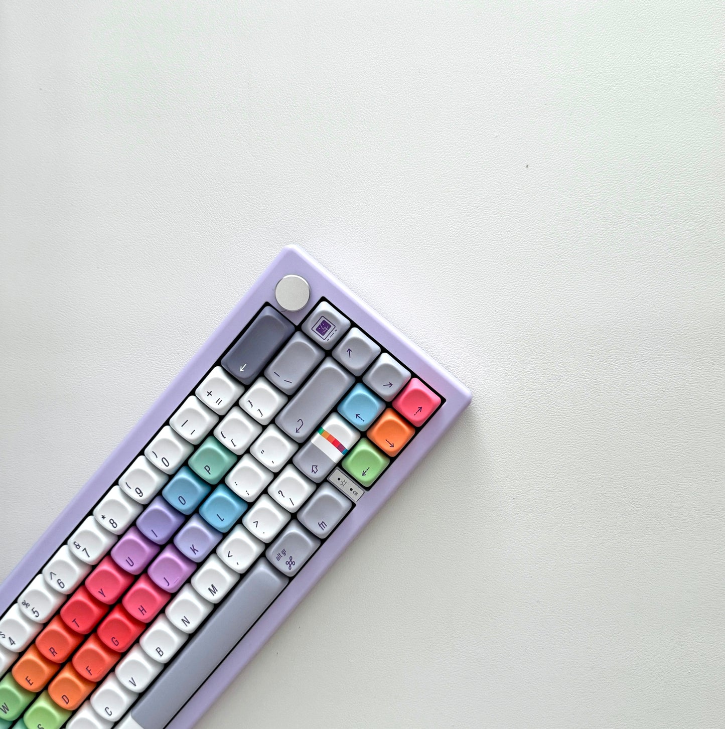 A compact mechanical keyboard for Mac with a pastel lavender frame and multicolor rainbow keycaps, photographed diagonally on a clean off-white surface.