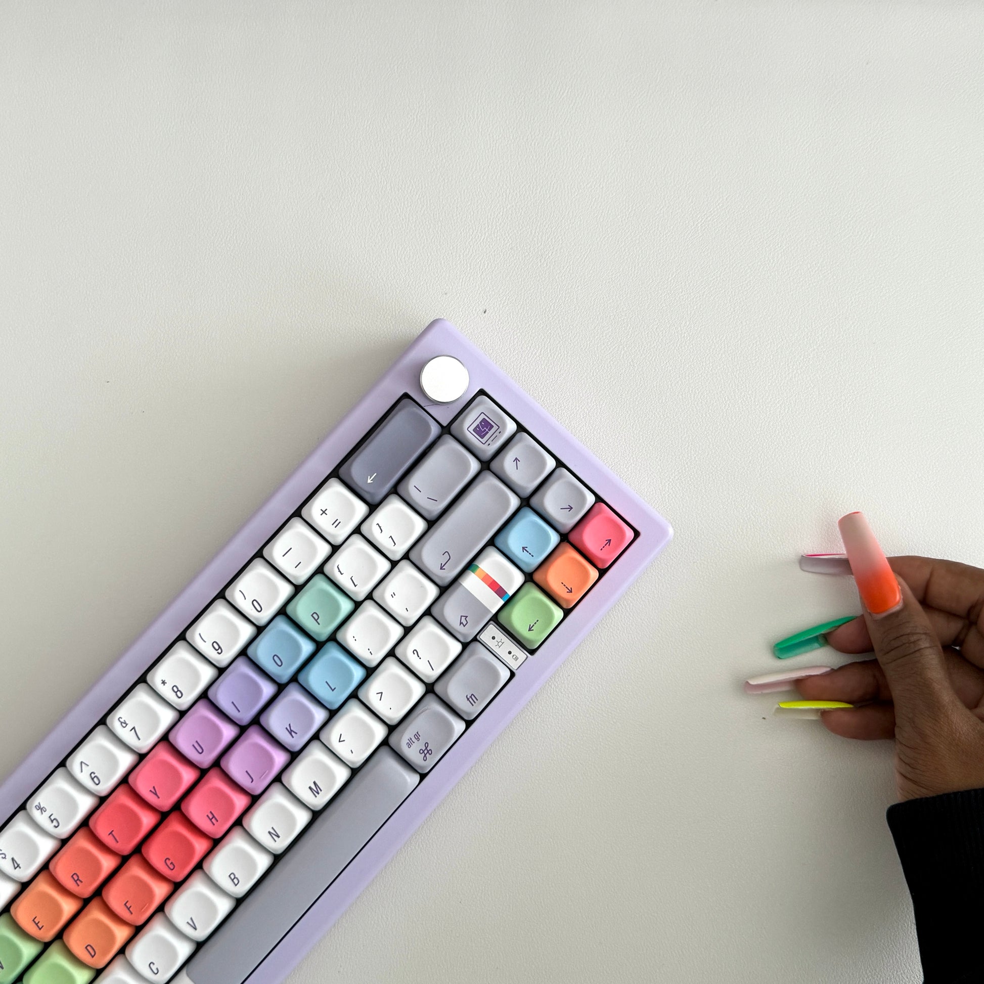 A pastel rainbow mechanical keyboard with a lavender-purple frame is shown from an overhead angle on a white surface, next to a hand with long orange, green, pink, and yellow nails holding three matching miniature keycap sets.