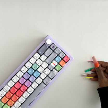 A pastel rainbow mechanical keyboard with a lavender-purple frame is shown from an overhead angle on a white surface, next to a hand with long orange, green, pink, and yellow nails holding three matching miniature keycap sets.